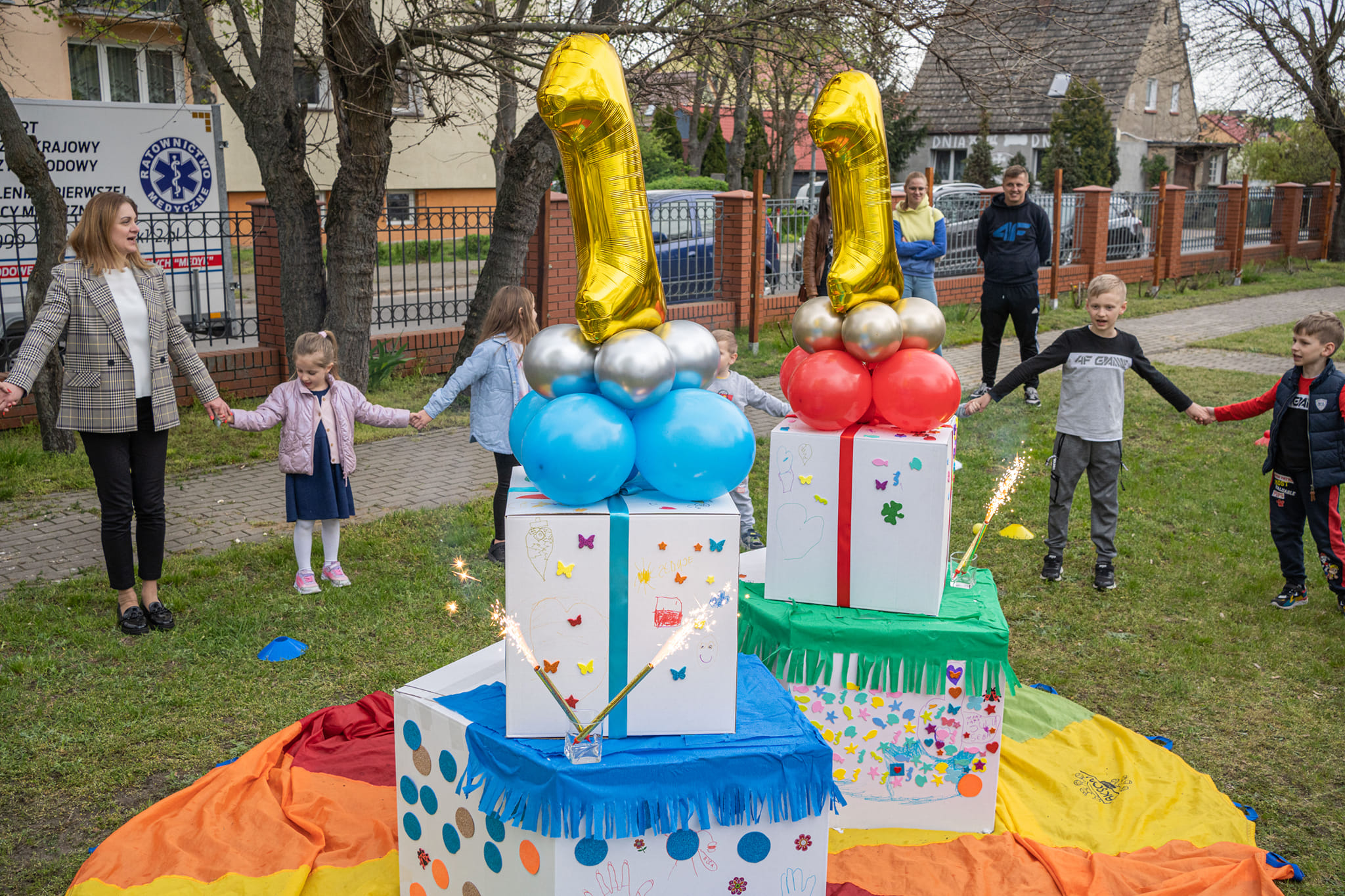 kartony imitujące tort, na wierzchu dwa duże złote balony w kształcie cyfry 1, wokół nich w kółku stoją ludzie, którzy trzymaja się za ręce